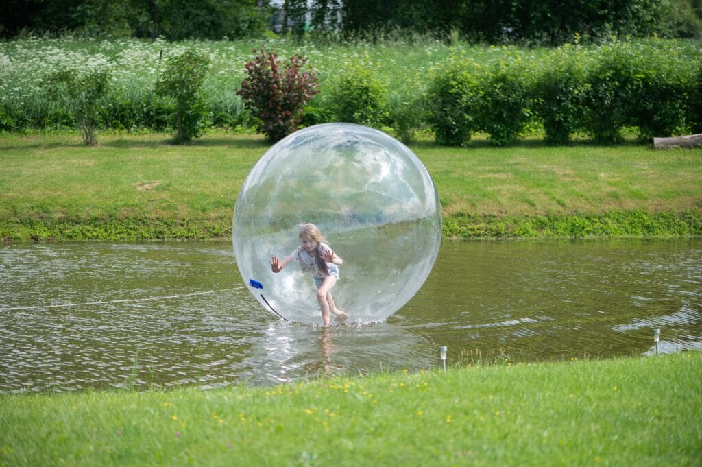 Child inside a large inflatable ball on a pond, surrounded by lush greenery, enjoying an outdoor activity.