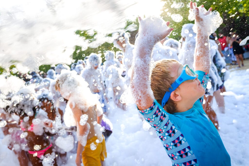 Children playing joyfully in a large foam pit at a party, surrounded by colorful decorations and bright sunlight.