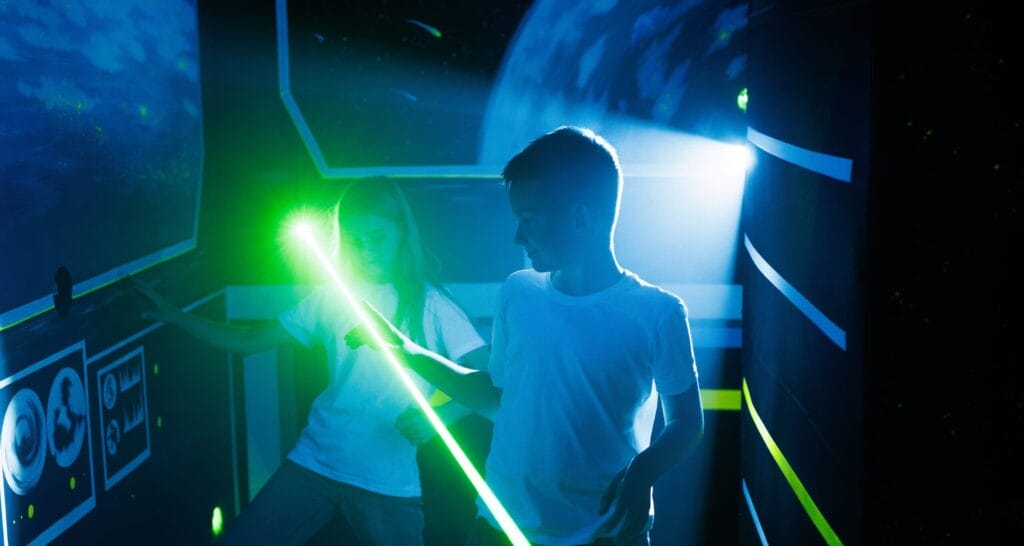 A girl and boy navigate a laser labyrinth simulation game, interacting with beams of light in a dark, immersive environment.
