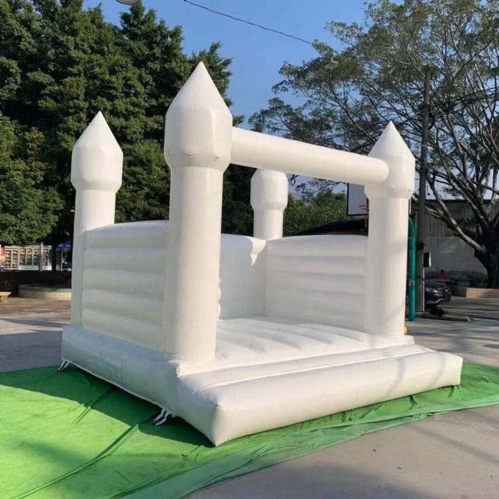 White bounce house set up outdoors on a sunny day, surrounded by green grass and trees in the background.